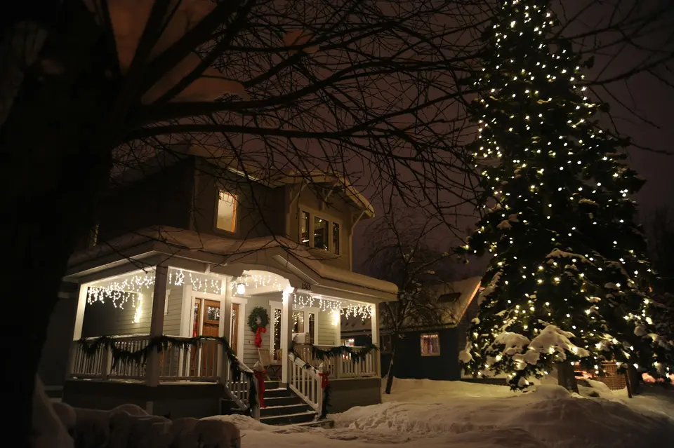 A snow-covered house at night with a porch trimmed in white lights and a tall spruce wrapped in warm white string lights